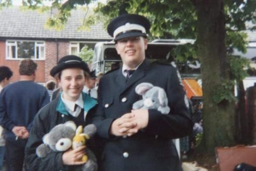 Colour photograph of two Cadets staning outside in front of a bus holding cuddly toys, wearing black and white St John uniform.