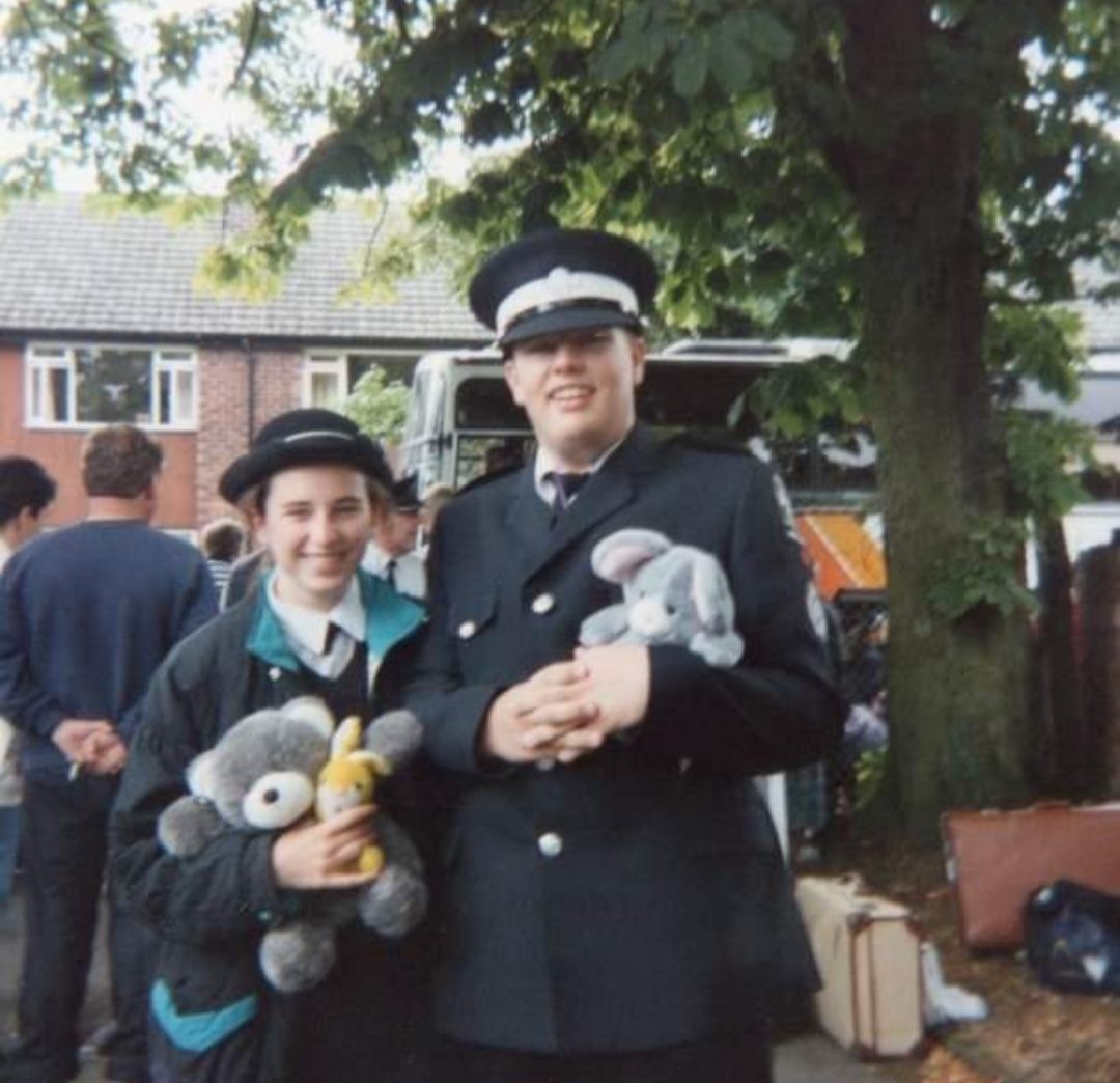 Colour photograph of two Cadets staning outside in front of a bus holding cuddly toys, wearing black and white St John uniform.