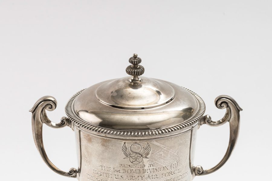 Silver trophy with lid and two handles standing on a wooden base covered with silver plaques bearing the names of former winners.