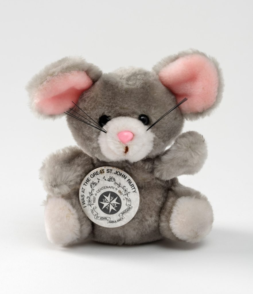 Colour photograph of a grey cuddly toy mouse with pink ears and black whiskers. It wears a badge reading 'I was at the Great St John Party' 1987'.
