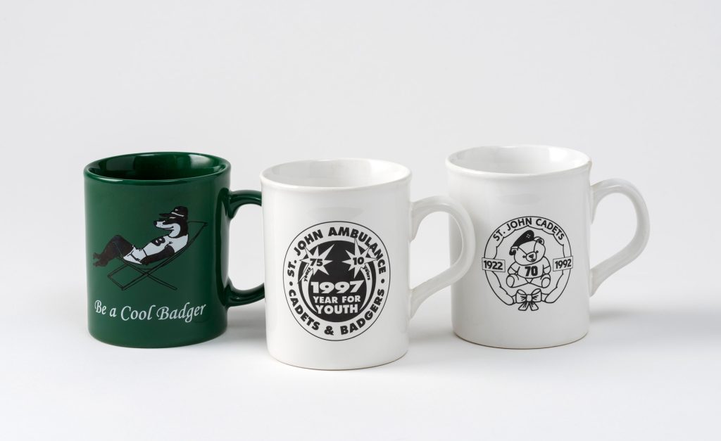 Colour photograph of three mugs, from left to right: a green mug with a Badger relaxing in a deckchair and inscribed 'Be a Cool Badger', a white and black mug celebrating the year of youth in 1997 and a black and white mug celebrating 70 years of Cadets in 1992