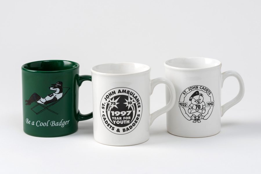 Colour photograph of three mugs, from left to right: a green mug with a Badger relaxing in a deckchair and inscribed 'Be a Cool Badger', a white and black mug celebrating the year of youth in 1997 and a black and white mug celebrating 70 years of Cadets in 1992