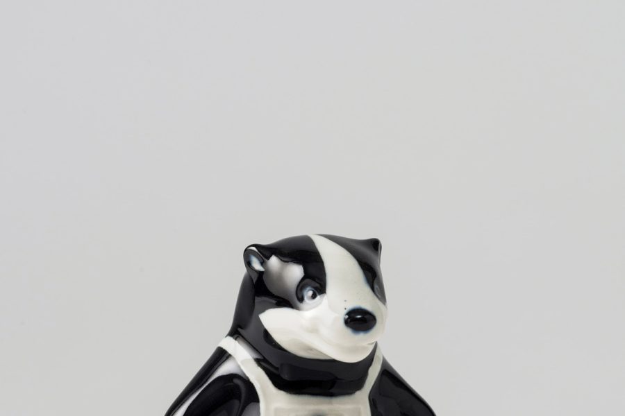 Colour photograph of a small black and white porcelain figurine of a badger wearing white sleeveless dungarees with a letter B in black on the front.