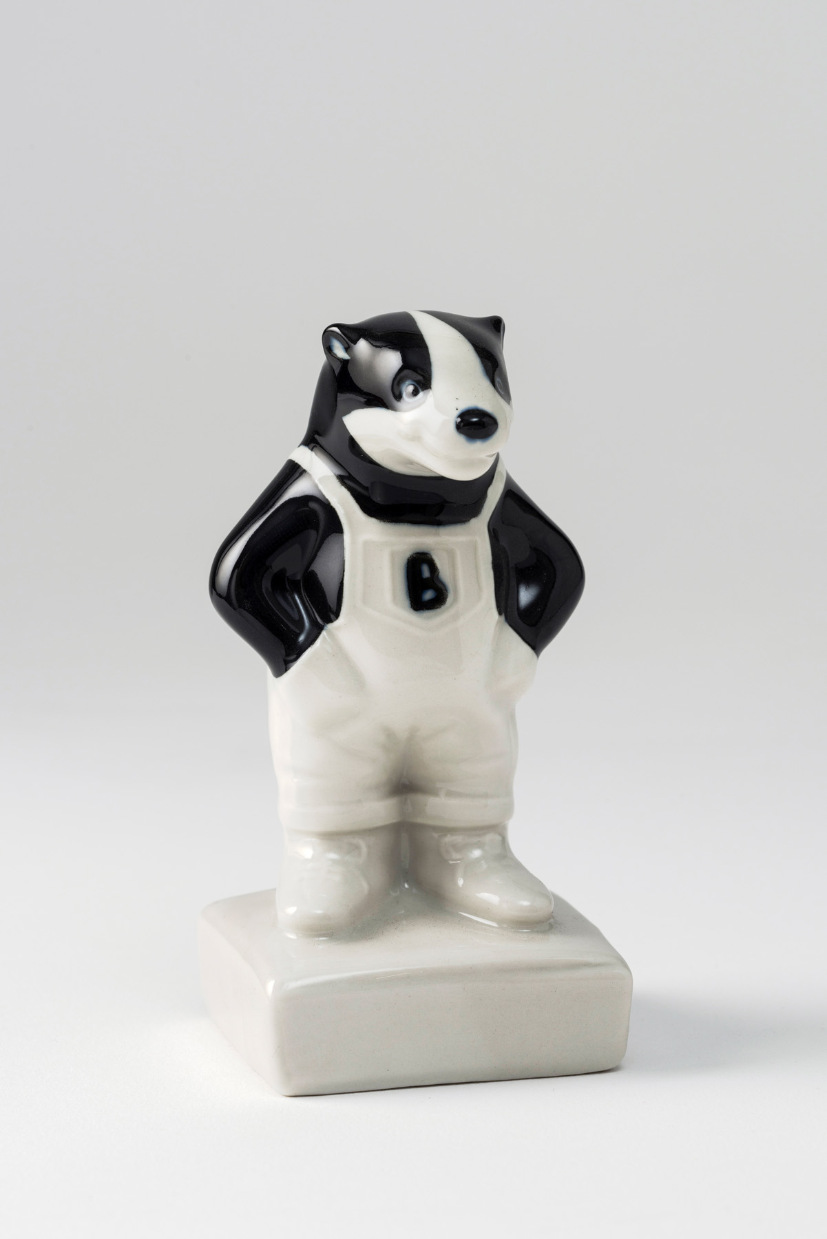 Colour photograph of a small black and white porcelain figurine of a badger wearing white sleeveless dungarees with a letter B in black on the front.