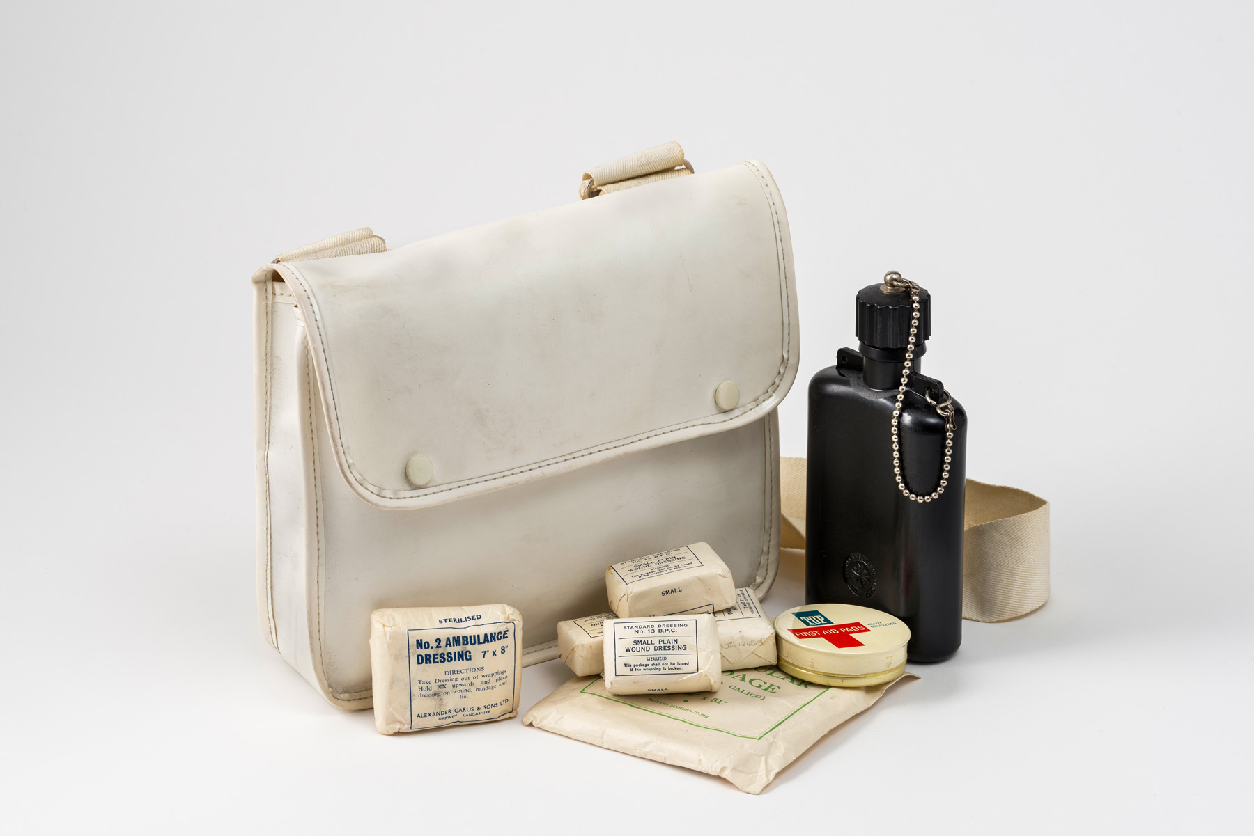 White rectangular shaped bag with two white poppers and a white canvas strap. The photograph shows the contents of the bag on display including a black plastic water bottle and various dressings and bandages.