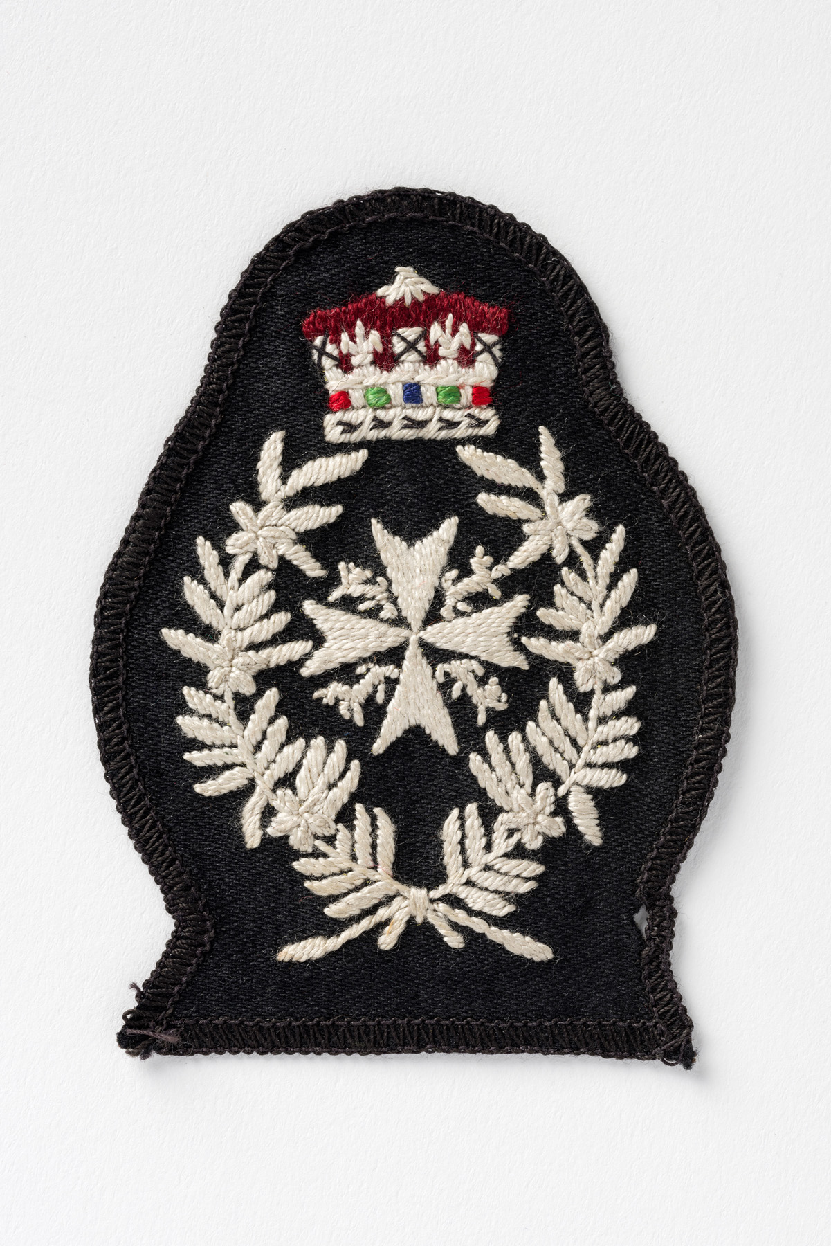 Colour photograph of a black cloth badge embroidered with the white eight-pointed of cross of St John, surmounted by a Crown and encircled by two branches of leaves