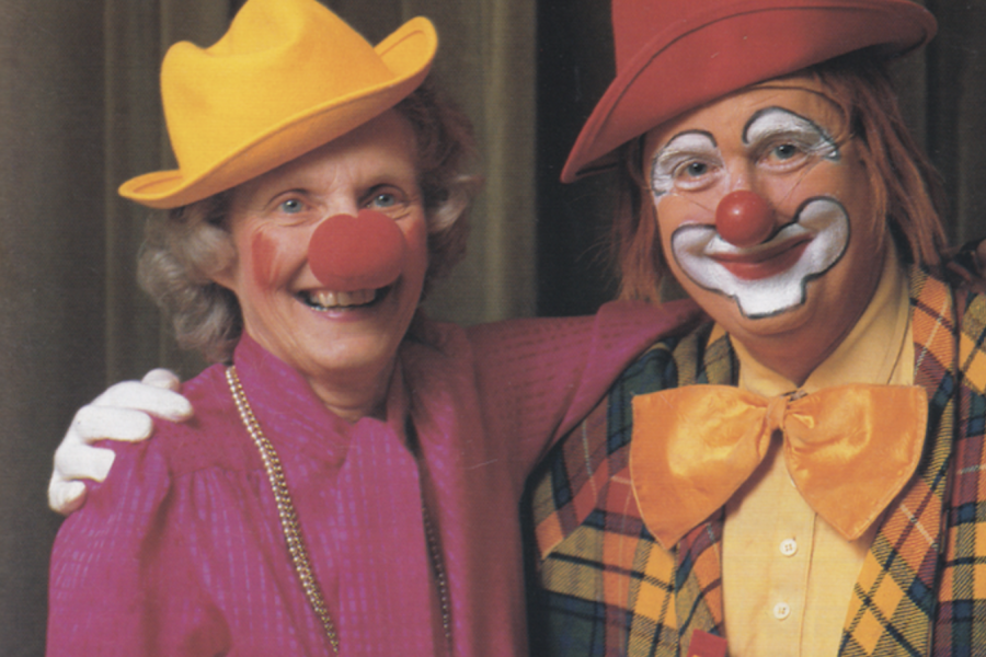 Colour photograph of a man and woman. The man on the right is dressed as a clown. The woman on the left is wearing a foam red nose.
