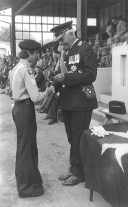 A boy is standing on the left, dressed in Cadet uniform. He is receiving a trophy shield from a uniformed adult, who is standing on the right. In the background are lots of people sitting on raised seating.