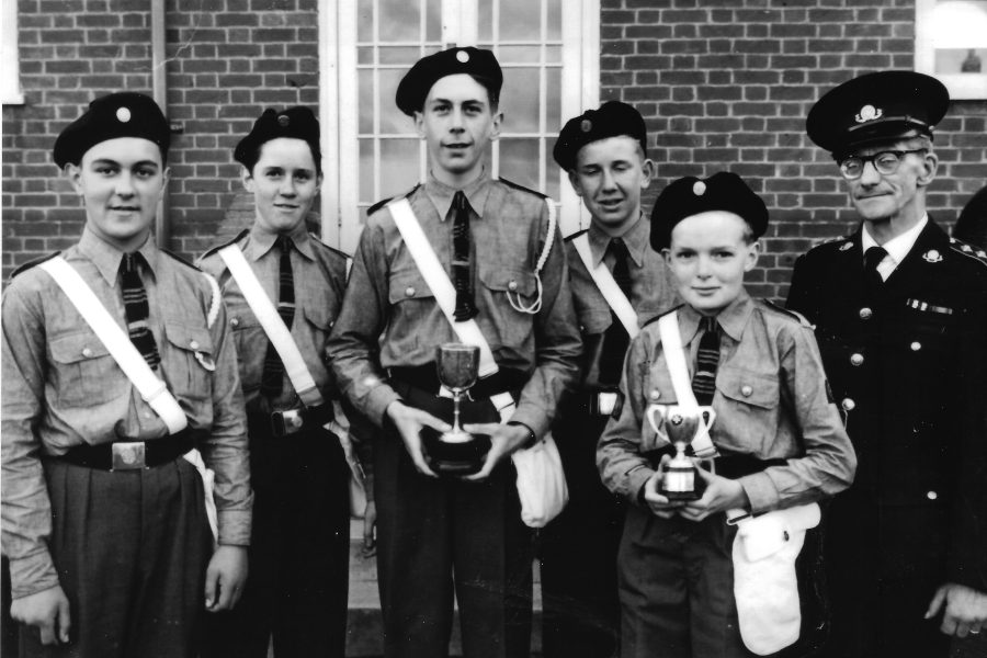 Black and white photograph of five male Cadets in uniform standing in a row. At the end of the row is an adult male in uniform. Two of the Cadets are holding trophies.