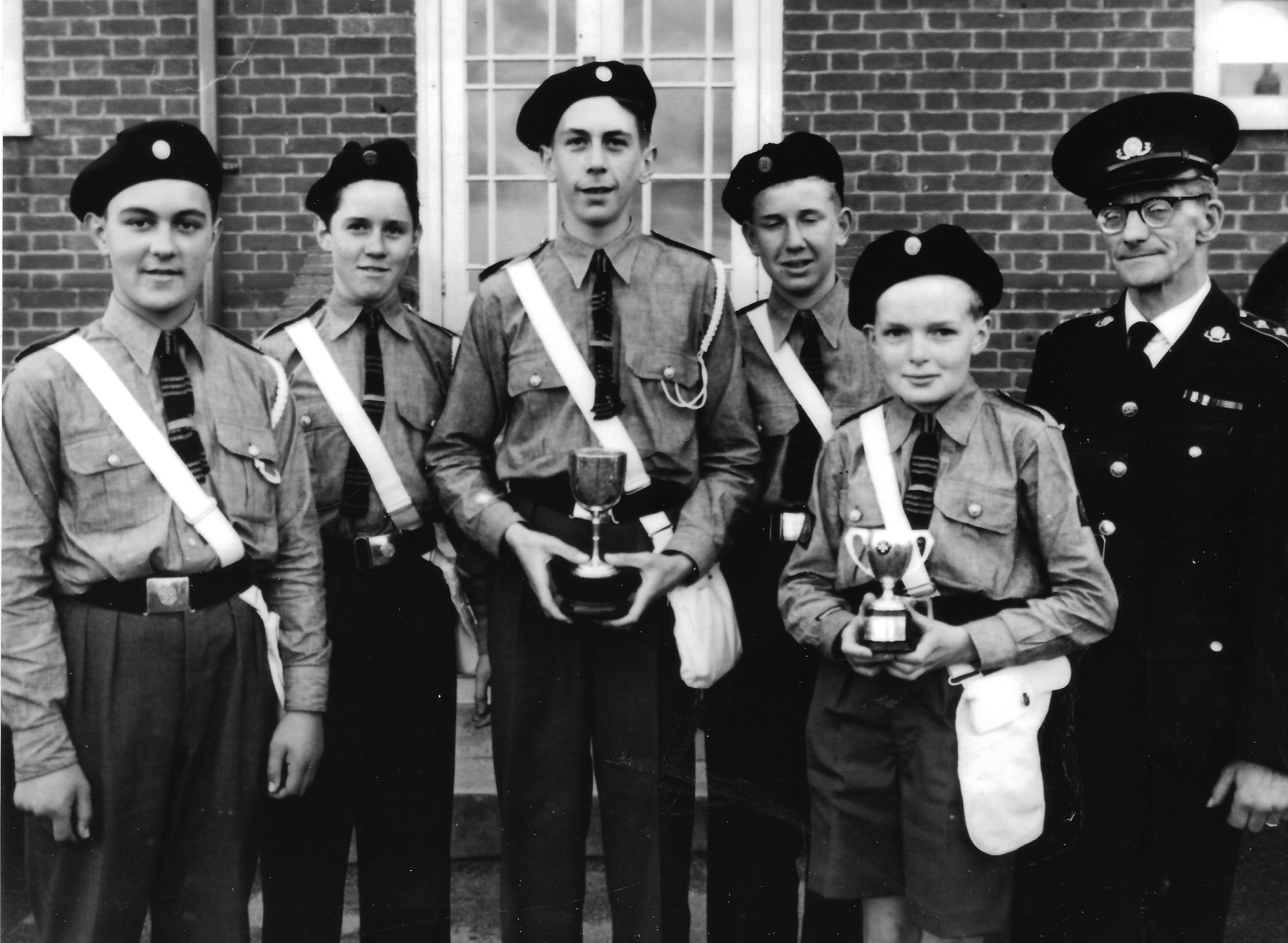 Black and white photograph of five male Cadets in uniform standing in a row. At the end of the row is an adult male in uniform. Two of the Cadets are holding trophies.